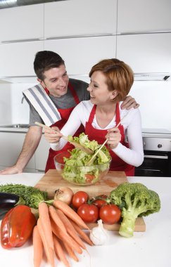 beautiful American couple working at home kitchen in apron mixing vegetable salad smiling happy