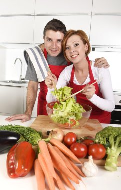 beautiful American couple working at home kitchen in apron mixing vegetable salad smiling happy