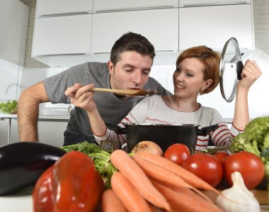 young attractive couple at home kitchen with man tasting vegetable stew cooked by her wife smiling happy