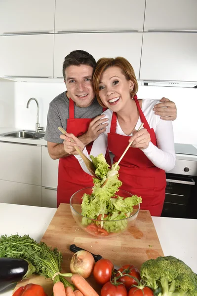 Beautiful American couple working at home kitchen in apron mixing vegetable salad smiling happy — Stok fotoğraf