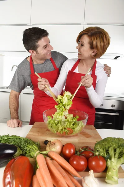 Beautiful American couple working at home kitchen in apron mixing vegetable salad smiling happy — Stockfoto