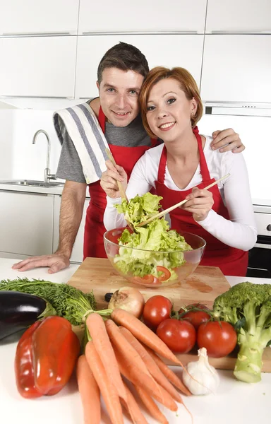 Beautiful American couple working at home kitchen in apron mixing vegetable salad smiling happy — Stockfoto