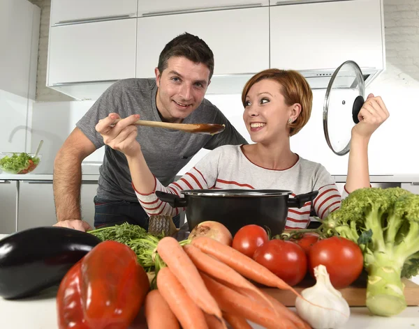 Joven atractiva pareja en casa cocina con hombre degustación vegetal guiso cocinado por su esposa sonriendo feliz —  Fotos de Stock