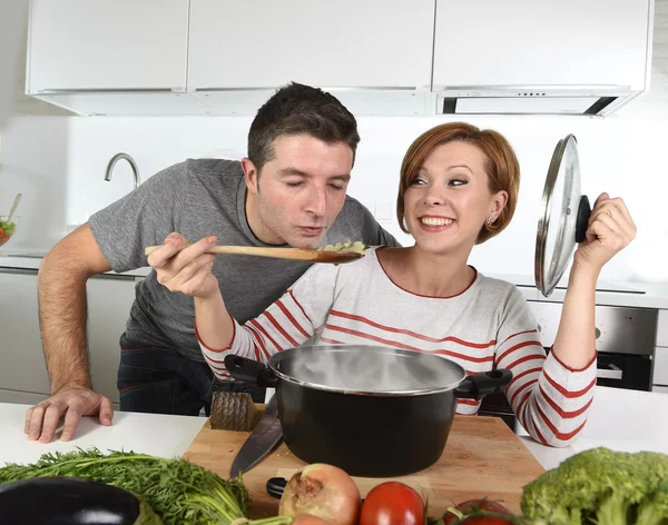 Joven atractiva pareja en casa cocina con hombre degustación vegetal guiso cocinado por su esposa sonriendo feliz —  Fotos de Stock