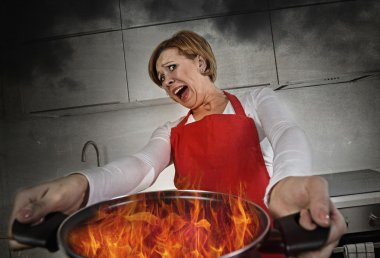 young inexperienced home cook woman in panic with apron holding pot burning in flames with in panic