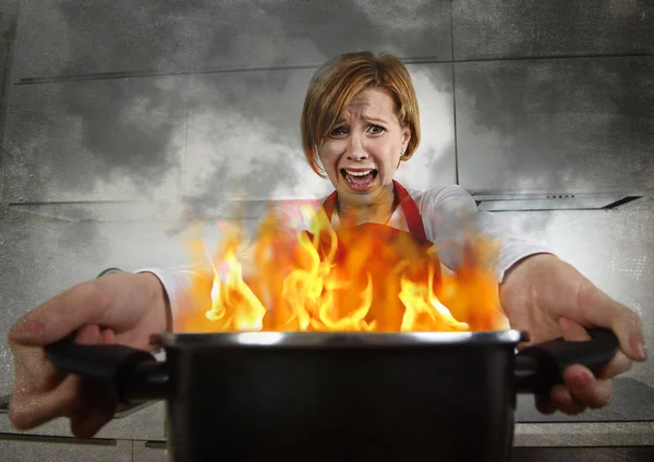 Young inexperienced home cook woman in panic with apron holding pot burning in flames with in panic — Stok fotoğraf