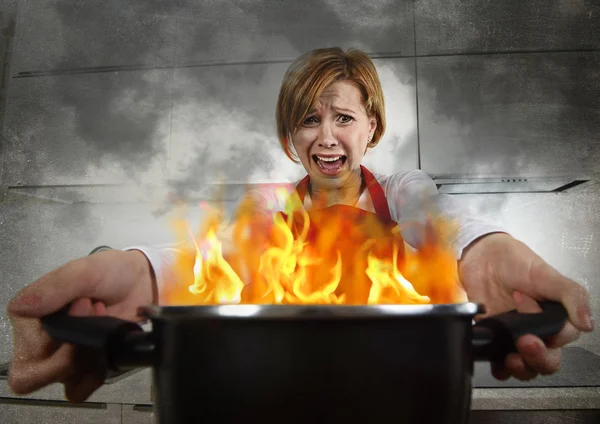 young inexperienced home cook woman in panic with apron holding pot burning in flames with in panic