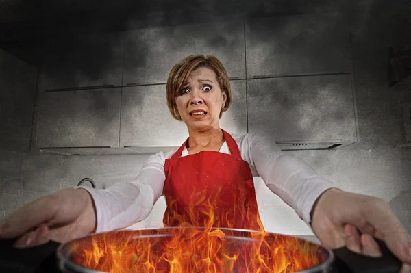 Young inexperienced home cook woman in panic with apron holding pot burning in flames with in panic — Stockfoto