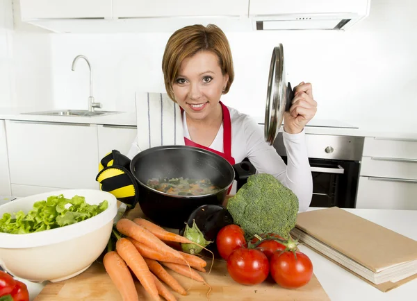 Домашний повар женщина в красном фартуке на домашней кухне держа кастрюлю с горячим супом нюхает овощное рагу — стоковое фото