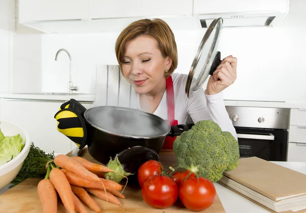 Home cook woman in red apron at domestic kitchen holding cooking pot with hot soup smelling vegetable stew — Stockfoto