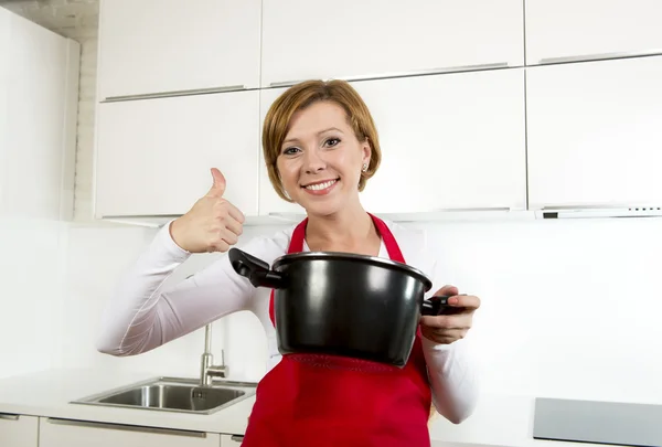 Young beautiful home cook woman in red apron at domestic kitchen holding spoon and cooking pot with delicious soup — Stockfoto