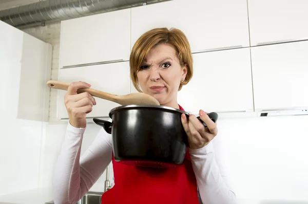 Home cook woman at kitchen holding cooking pot and spoon tasting soup in a funny disgusting bad taste face — Stockfoto