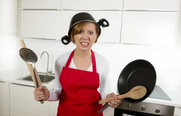Junge attraktive Hausfrau in roter Schürze in der Küche hält Pfanne und Haushalt mit Topf auf dem Kopf im Stress — Stockfoto