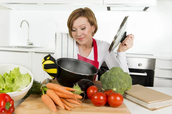 Home cook woman in red apron at domestic kitchen holding cooking pot with hot soup smelling vegetable stew — Stockfoto