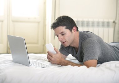 young attractive man lying on bed or couch using mobile phone and computer laptop working from home