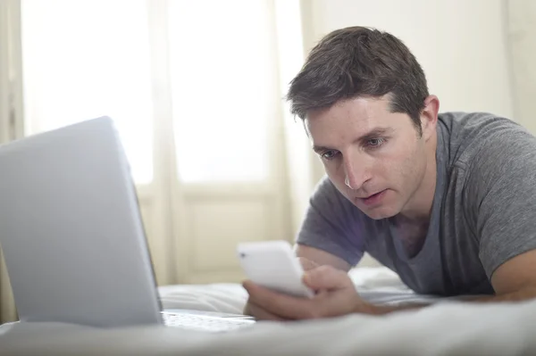 Young attractive man lying on bed or couch using mobile phone and computer laptop internet addict — Stockfoto