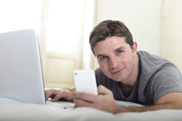Jovem atraente homem deitado na cama ou no sofá usando telefone celular e computador laptop viciado em internet — Fotografia de Stock
