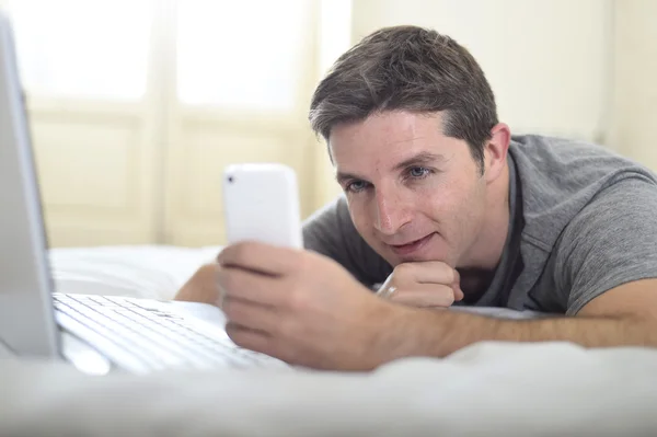Joven atractivo hombre acostado en la cama o sofá utilizando el teléfono móvil y ordenador portátil adicto a Internet —  Fotos de Stock
