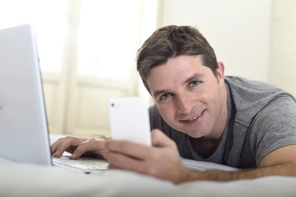 Young attractive man lying on bed or couch using mobile phone and computer laptop internet addict — Stock Photo, Image