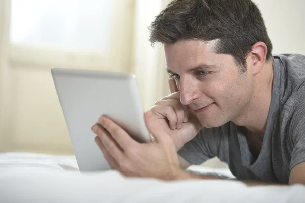 Молодой привлекательный мужчина лежит на кровати или диване наслаждаясь социальными сетями с помощью цифрового планшетного компьютера Интернет дома — стоковое фото