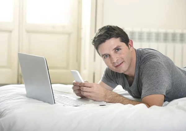 Jovem homem atraente deitado na cama ou no sofá usando telefone celular e laptop de computador trabalhando em casa — Fotografia de Stock