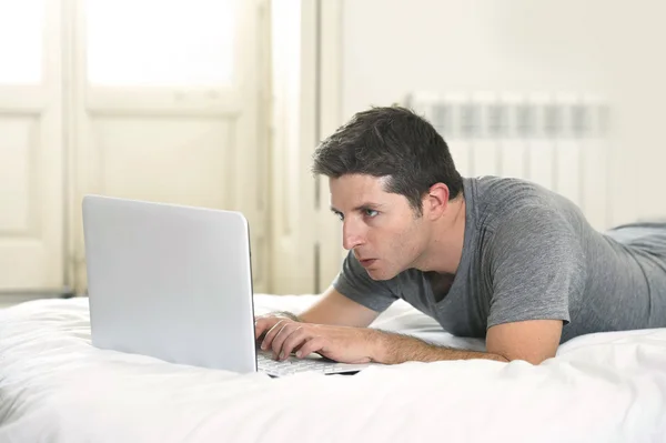 Junger attraktiver Mann, der auf Bett oder Couch liegt und am Laptop tippt, der mit dem Internet verbunden ist — Stockfoto