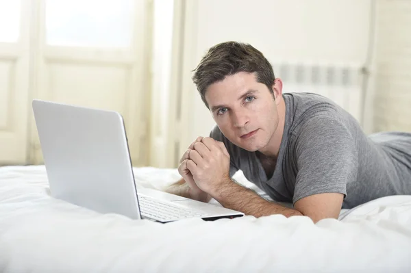 Jovem homem atraente deitado na cama ou no sofá desfrutando de redes sociais usando laptop computador em casa internet sem fio — Fotografia de Stock