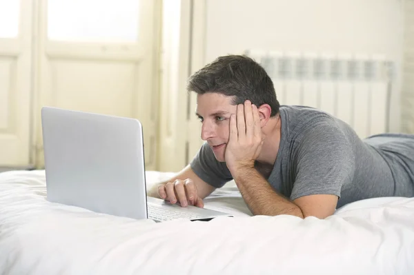 Young attractive man lying on bed or couch enjoying social networking using computer laptop at home — ストック写真