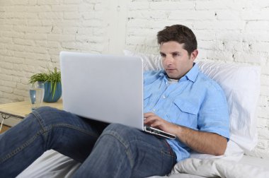 young attractive man lying on bed enjoying social networking using computer laptop at home