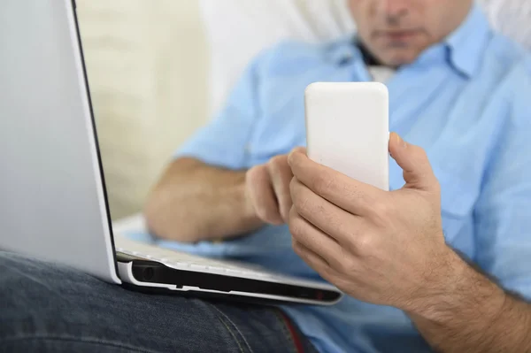 Cerrar las manos del hombre acostado en el sofá de la cama usando el teléfono móvil y la computadora que trabaja desde casa — Foto de Stock