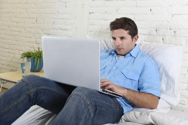 Young attractive man lying on bed enjoying social networking using computer laptop at home — ストック写真