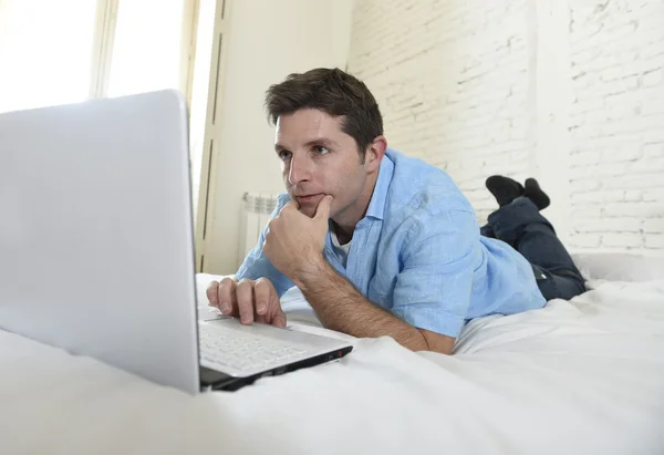 Jovem atraente homem deitado na cama desfrutando de redes sociais usando computador em casa — Fotografia de Stock