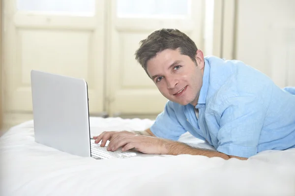 Jovem atraente homem deitado na cama desfrutando de redes sociais usando computador em casa — Fotografia de Stock