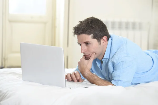 Jovem homem atraente deitado na cama desfrutando de redes sociais usando laptop em casa — Fotografia de Stock