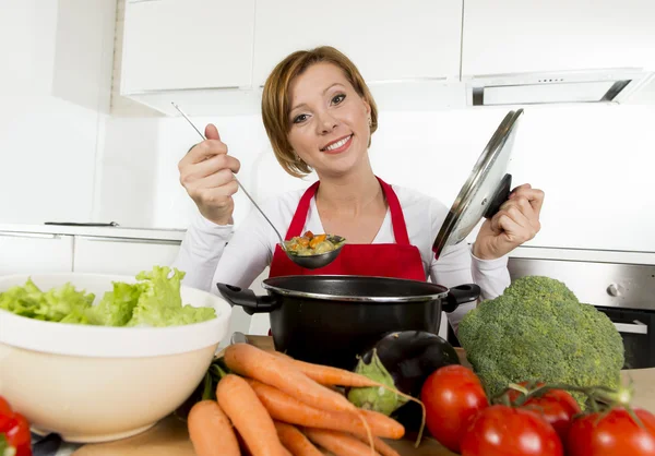 Young happy home cook woman in red apron at domestic kitchen holding saucepan tasting hot soup — Stockfoto