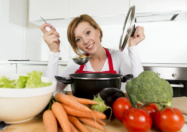 Young happy home cook woman in red apron at domestic kitchen holding saucepan tasting hot soup — Stockfoto