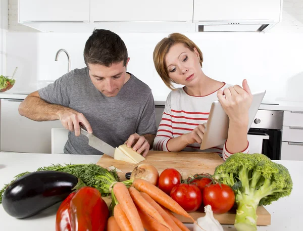 Pareja americana en la cocina doméstica esposa siguiendo la receta en almohadilla digital trabajando junto con el marido —  Fotos de Stock