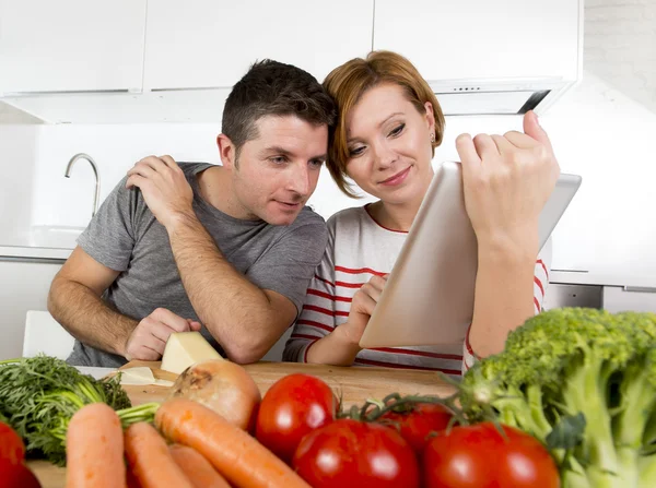 Pareja americana en la cocina doméstica esposa siguiendo la receta en almohadilla digital trabajando junto con el marido —  Fotos de Stock