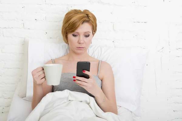 Having coffe breakfast on bed while watching internet news in his mobile phone in online communication — Stockfoto