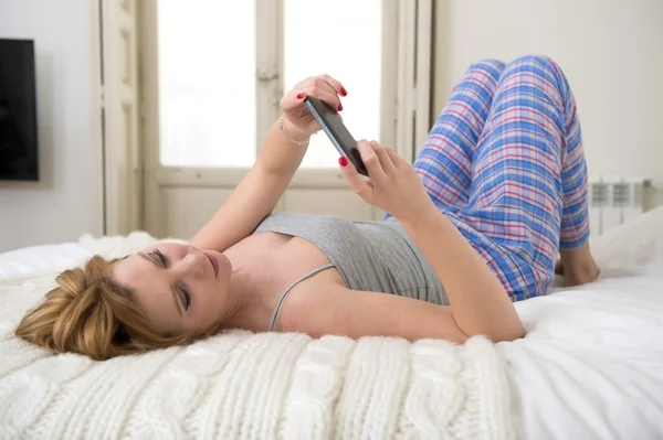 Chica atractiva joven con el pelo rojo de red de Internet con teléfono móvil tumbado en la cama en los pantalones de dormir —  Fotos de Stock