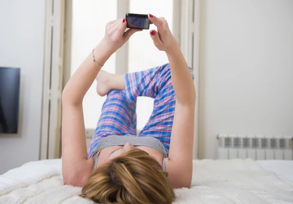 Young attractive girl with red hair internet networking with mobile phone lying on bed in sleeping pants — Φωτογραφία Αρχείου