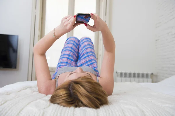 Jovem menina atraente com cabelo vermelho internet networking com telefone móvel deitado na cama em calças de dormir — Fotografia de Stock