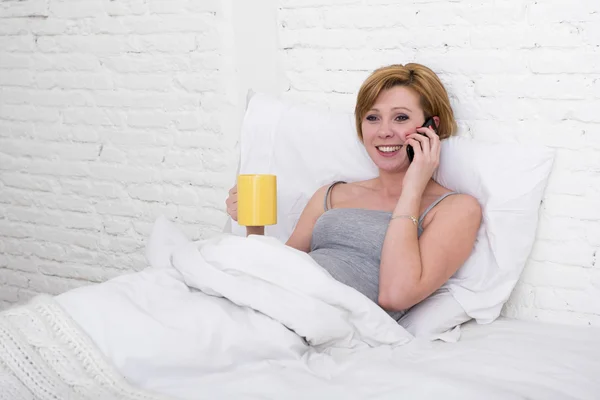 Young beautiful girl having coffe on bed while watching internet news in his mobile phone in online communication — Stok fotoğraf