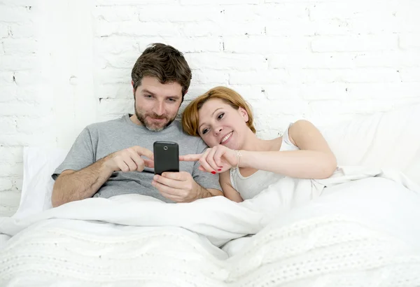 Счастливая привлекательная пара в постели с помощью мобильного телефона улыбаясь смотреть вместе интернет-приложение — стоковое фото