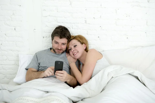 Feliz pareja atractiva en la cama usando el teléfono móvil sonriendo viendo juntos aplicación de Internet —  Fotos de Stock