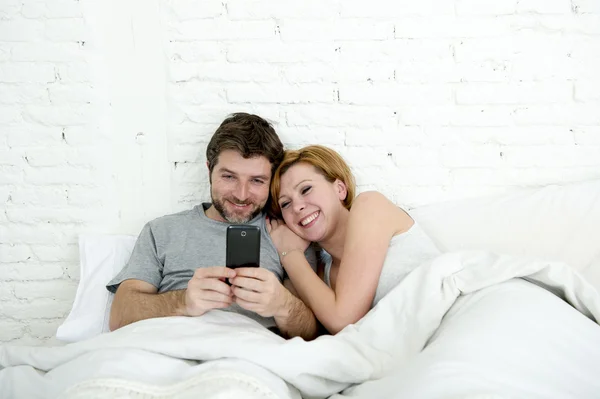 Happy attractive couple in bed using mobile phone smiling watching together internet app — Zdjęcie stockowe