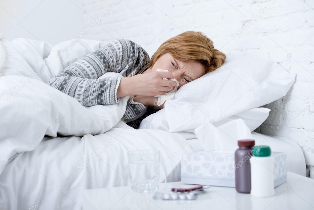 woman with sneezing nose blowing in tissue on bed suffering cold flu virus symptoms having medicines tablets pills