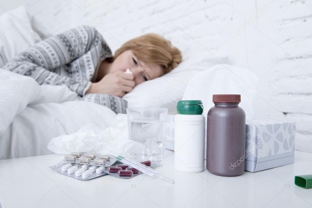 woman with sneezing nose blowing in tissue on bed suffering cold flu virus symptoms having medicines tablets pills