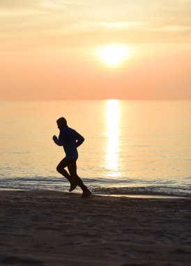 Siluet genç adam turuncu gökyüzüyle gün batımında sahilde koşuyor.