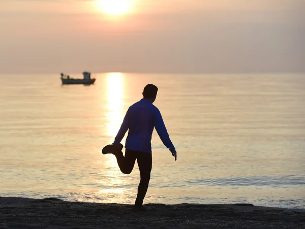 Silhouette eines jungen Sportlers, der nach dem Lauftraining im Freien am Strand bei Sonnenuntergang ein Bein streckt — Stockfoto